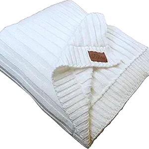 Puede incluir: Una manta de punto plegada, de color blanco roto, con un diseño acanalado. La manta tiene forma rectangular y una etiqueta de cuero marrón con un logotipo. Fabricada con un material suave y texturizado, ideal para la decoración del hogar.