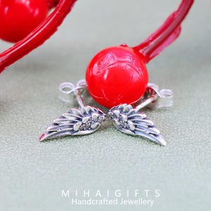 May include: A pair of silver angel wing earrings with small clear cubic zirconia stones. The earrings are on a green surface with a red ball in the background.
