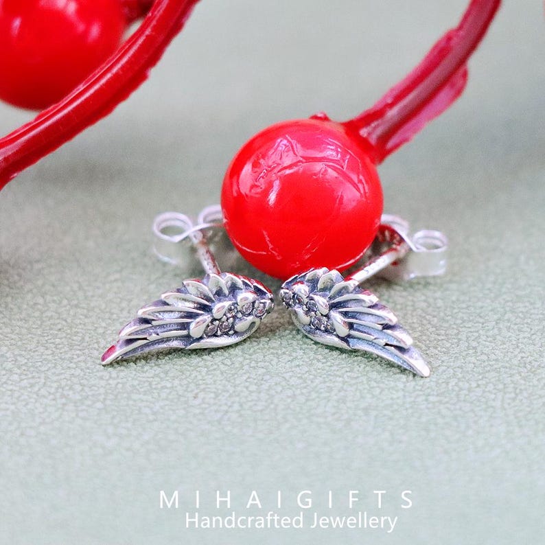 May include: A pair of silver angel wing earrings with small clear cubic zirconia stones. The earrings are on a green surface with a red ball in the background.