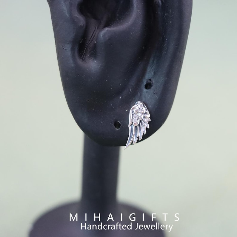 May include: Silver angel wing earring with clear cubic zirconia stones. The earring is shown on a black mannequin ear.