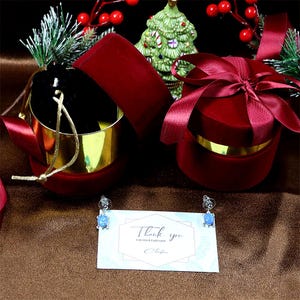 May include: Two red velvet gift boxes with gold trim, one with a satin ribbon, and a pair of blue opal earrings on a thank you card.