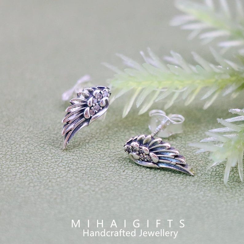 May include: A pair of silver earrings with a wing design and small clear stones. The earrings are on a green background.