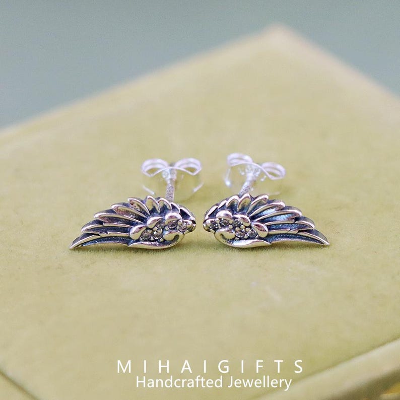 May include: A pair of silver angel wing earrings with small clear cubic zirconia stones. The earrings are on a green velvet background.