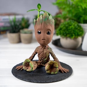 Puede incluir: Figura marrón, similar a Groot, sentado con un brote que crece de su cabeza. La figura tiene ojos grandes y oscuros y está sobre una base redonda negra. Es un objeto decorativo, posiblemente una maceta o un objeto de colección.