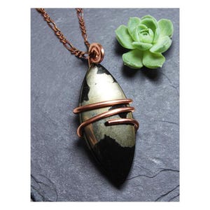May include: A handcrafted pendant necklace featuring a polished, elongated stone with a metallic sheen, wrapped in copper wire. The pendant hangs from a copper-colored chain. A small green succulent is in the background.