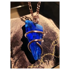 May include: A vibrant blue crystal pendant necklace. The crystal is wrapped with copper wire and hangs from a copper chain. The pendant has a unique, irregular shape, and the chain is a classic link style. The necklace is displayed on a dark, textured surface.
