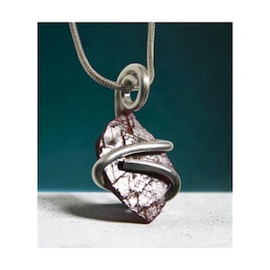 May include: A silver pendant necklace featuring a rough, dark red gemstone wrapped in a silver wire design. The pendant hangs from a silver chain, creating a striking contrast against the stone's texture and color. The background is a gradient of teal and white.