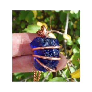 May include: A raw, deep blue crystal pendant wrapped in copper wire. The pendant is suspended from a copper chain. The background is a blurred view of green foliage.