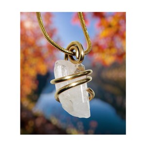 May include: A gold-tone necklace with a clear quartz crystal pendant. The crystal is encased in gold wire and hangs from a gold chain. The backdrop is a blurred image of autumn leaves and a lake.