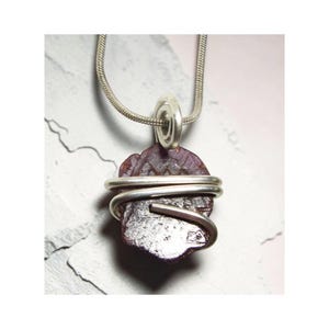 Puede incluir: Un collar con colgante artesanal con una gema en bruto de color púrpura oscuro envuelta en alambre plateado. El colgante cuelga de una delicada cadena plateada. La gema tiene una superficie rugosa y texturizada.