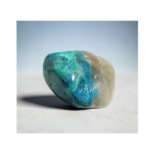 May include: A polished stone with a mix of colours. The stone has a smooth, rounded shape. The colours include shades of blue and teal, with a light grey area. The stone is on a light grey surface.