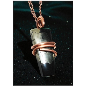 Op de afbeelding: Een rechthoekige hangerketting met een koperen ketting en draadwikkeling. De hanger is een donkere, rechthoekige steen met zilver- en zwarttinten. De koperdraad is om de steen gewikkeld. De achtergrond is donker en glinsterend. De steen is ongeveer 3 cm lang.