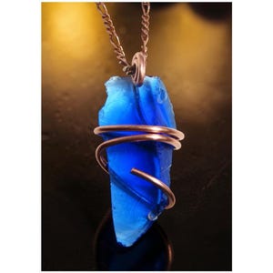 May include: A vibrant blue glass pendant, irregularly shaped, is suspended from a delicate copper-colored chain. The glass is wrapped with copper wire. The pendant is a striking piece of jewelry.