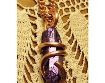 Op de afbeelding: Een koperkleurige ketting met een paarse kristallen hanger. Het kristal is omwikkeld met koperdraad en hangt aan een bijpassende ketting. De achtergrond is een crèmekleurige stof met open weefsel.