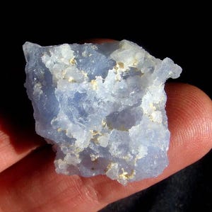 May include: A raw, translucent blue chalcedony mineral specimen with a jagged, crystalline texture. The stone features a pale, icy blue coloration with scattered white, opaque inclusions across its uneven, rough surface and edges.