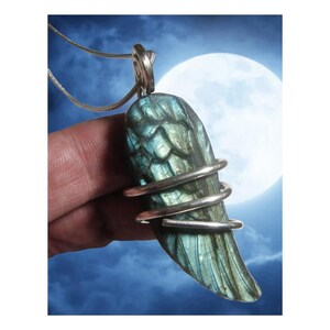 May include: A silver pendant featuring a blue-green labradorite gemstone carved into the shape of an angel wing. The gemstone is wrapped with silver wire.