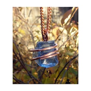 May include: A blue crystal pendant wrapped in copper wire, suspended from a copper chain. The crystal has a rough, natural texture. The background is blurred with autumn foliage.