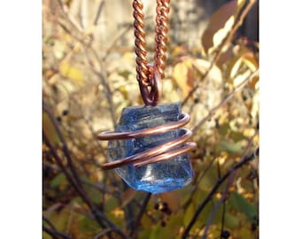 May include: A blue crystal pendant wrapped in copper wire, suspended from a copper chain. The crystal has a rough, natural texture. The background is blurred with autumn foliage.