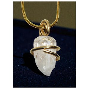 May include: A gold necklace with a clear crystal pendant. The crystal is encircled by a gold band and hangs from a gold chain. The pendant has a natural, irregular shape, and the chain has a fine, textured design.