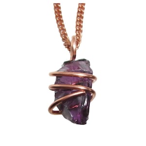 May include: A raw, purple amethyst pendant wrapped in copper wire, suspended from a copper chain. The amethyst has a rough, natural texture, and the wire is coiled around the stone. The chain is a twisted rope design.
