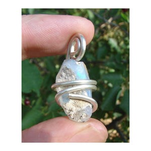 Peut inclure: Pendentif en opale brute enveloppé dans du fil d'argent. L'opale a une couleur bleu clair et irisée. Le fil est enroulé autour de la pierre et forme une boucle en haut pour l'accrocher. Le pendentif est tenu par un doigt.