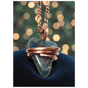 May include: A handmade pendant with a blue-green stone encased in copper wire, suspended from a copper chain. The pendant is set against a blurred backdrop of warm, out-of-focus lights.