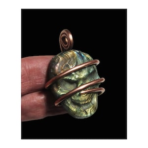 May include: A skull-shaped pendant with a green and gold iridescent finish, wrapped with copper wire. The pendant has a copper spiral at the top for hanging. The skull is held in a person's hand against a black background.