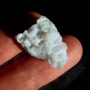 May include: A small, raw, translucent light blue chalcedony mineral specimen with a jagged, uneven surface texture. The stone displays a pale, icy blue coloration with subtle variations in opacity and crystalline structure across its irregular form.