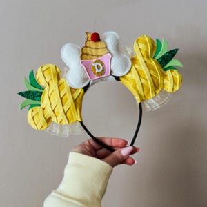 May include: A yellow and green headband with Mickey Mouse ears, designed to resemble a pineapple. The headband features a cupcake with a cherry on top and a pink container with the letter "D".