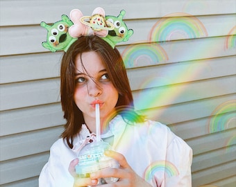 May include: A young person wearing a pink shirt and a headband with green alien ears and a pink and yellow spaceship design. They are holding a clear plastic cup with a straw and a green alien character on the side. The background is a light brown wall with rainbow designs.