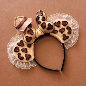 Peut inclure: Un serre-tête sur le thème du safari, de couleur beige et marron, avec un bandeau noir. Le serre-tête comprend un chapeau de safari, un nœud avec des taches de paillettes marron et des oreilles avec des taches de paillettes marron. Une bordure en dentelle blanche borde les oreilles.
