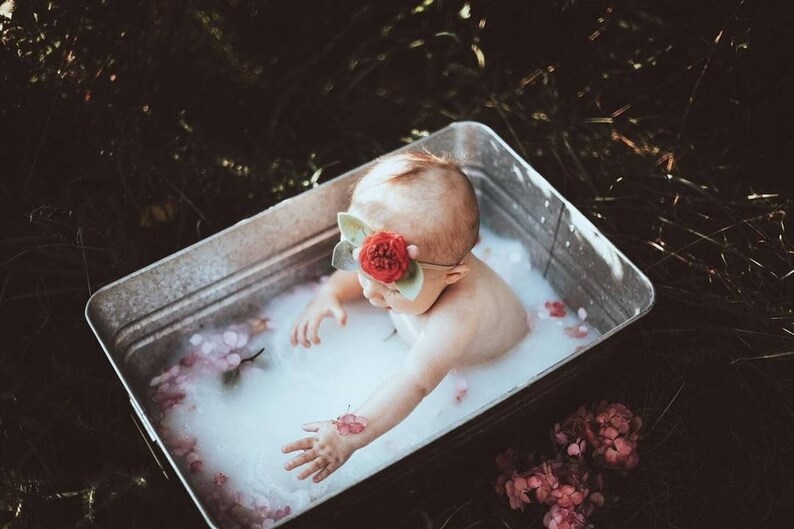 May include: A baby in a galvanized metal tub filled with milky water and flower petals. The baby wears a red flower headband. The scene is set outdoors with dark green grass and foliage.