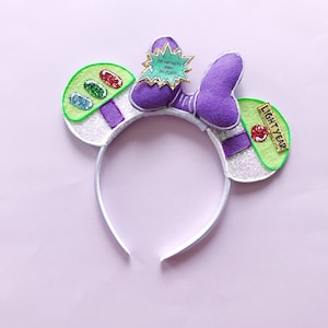 May include: White headband with purple and green felt ears. The ears feature a green space ranger design with a red button, a green, red, blue and yellow light, and the text "TO INFINITY AND BEYOND". The right ear has a green felt tag with the text "LIGHTYEAR".