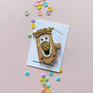 May include: A felt hair clip with a cartoon face. The face is brown with a gold glitter nose and mouth. The clip is attached to a brown felt rectangle with the text "accessories for children www.shopofrainbow.etsy.com" and "add and grow".