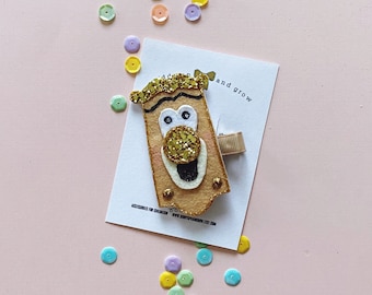 Op de afbeelding: Een haarspeld van vilt met een cartoon gezicht. Het gezicht is bruin met een gouden glitterneus en -mond. De speld is bevestigd aan een bruin vilt rechthoek met de tekst "accessoires for children www.shopofrainbow.etsy.com" en "add and grow".