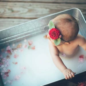 May include: A baby in a galvanized metal tub filled with milky water and flower petals. The baby wears a headband with a red rose and green leaves. The tub is on a wooden surface.