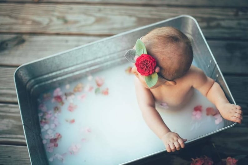 May include: A baby in a galvanized metal tub filled with milky water and flower petals. The baby wears a headband with a red rose and green leaves. The tub is on a wooden surface.