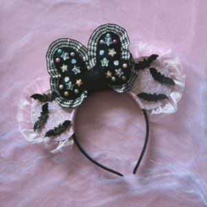 May include: A headband featuring two round ears covered in light pink glitter and black bat silhouettes. A large central bow is made of black fabric with a white gingham ruffle trim, decorated with various colorful star and circular beads.