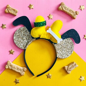 May include: A black headband with gold glitter ears and a yellow felt bow with a white bone and a green band. The headband is surrounded by small gold stars and dog bone-shaped biscuits with the text "Milk-Bone" on them.