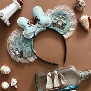May include: A nautical-themed headband with light blue Mickey Mouse ears, decorated with a brown anchor and a ship illustration. The headband has a black band and is decorated with white lace and a map design. Seashells and a ship in a bottle are also present.