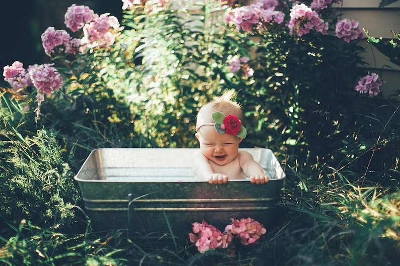 May include: A baby wearing a floral headband sits in a galvanized metal tub filled with water. The baby is surrounded by greenery and pink flowers, with a happy expression. The tub is outdoors.