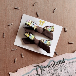 May include: Two brown velvet hair bows with a star and flower design. The bows are decorated with iridescent sequins and are displayed on a white card with the text "Ready Up and Grow".