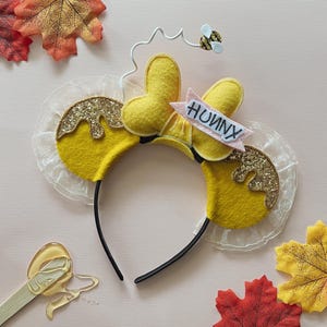 Peut inclure: Un serre-tête en feutre jaune avec des oreilles de souris, décoré de paillettes dorées et d'une bannière rose avec l'inscription "HUNNY". Une petite abeille en feutre est posée au-dessus des oreilles. Feuilles d'automne en arrière-plan.