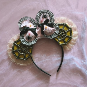 May include: A headband featuring two ears decorated with black and white gingham ruffles, yellow spider web patches, black bows, small plastic spiders, glittery bat cutouts, and pink beads. The base is a thin black band with sheer beige ruffles.
