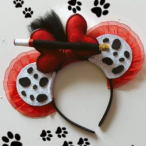 May include: Red and white dalmatian dog ears headband with a black and white striped wand. The headband is black and the ears are made of red fabric with white dalmatian spots. The wand is attached to the headband with a red bow.