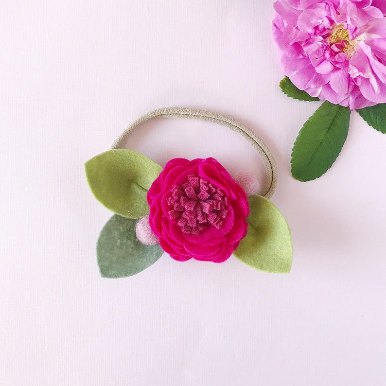 May include: A handmade hair tie featuring a vibrant fuchsia felt flower with a darker center, surrounded by two light green felt leaves. The elastic band is a neutral beige color. A pink flower is in the background.