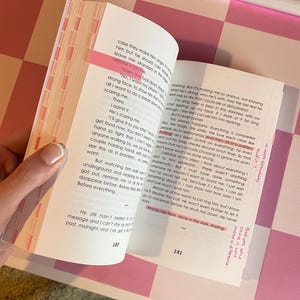 May include: An open book with pink page markers, displaying text on white pages. The book is held by a hand. The background features a pink and white chequered pattern and a keyboard.