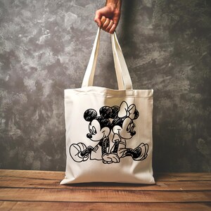 Pode incluir: Saco tote creme com um esbo&ccedil;o a preto e branco de Mickey e Minnie Mouse sentados juntos. O saco tem al&ccedil;as compridas e &eacute; segurado por uma m&atilde;o. O fundo &eacute; uma parede cinzenta e uma superf&iacute;cie de madeira.