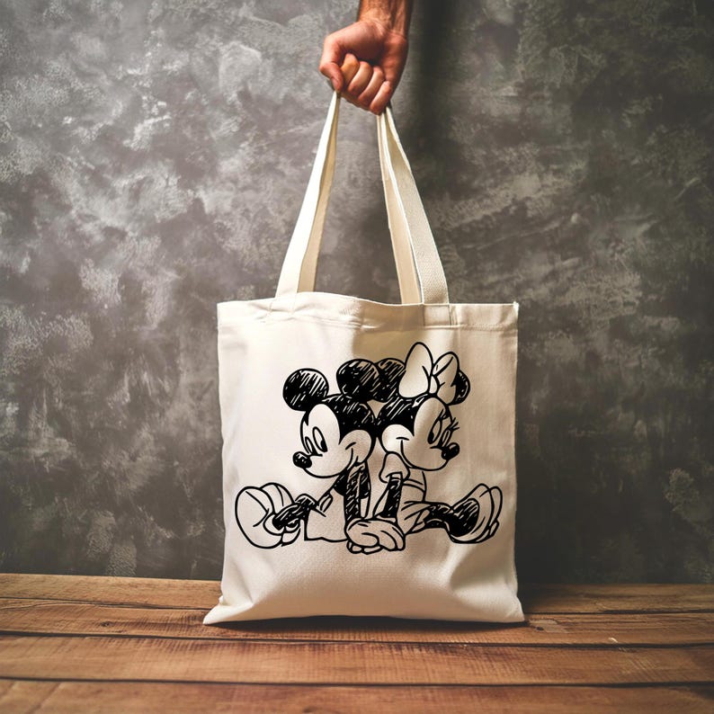 Pode incluir: Saco tote creme com um esbo&ccedil;o a preto e branco de Mickey e Minnie Mouse sentados juntos. O saco tem al&ccedil;as compridas e &eacute; segurado por uma m&atilde;o. O fundo &eacute; uma parede cinzenta e uma superf&iacute;cie de madeira.