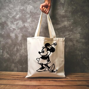 Pode incluir: Saco tote creme com uma ilustra&ccedil;&atilde;o a preto e branco da Minnie Mouse. O saco &eacute; segurado por uma m&atilde;o e tem al&ccedil;as compridas. O gr&aacute;fico da Minnie Mouse &eacute; um design cl&aacute;ssico de desenho animado.